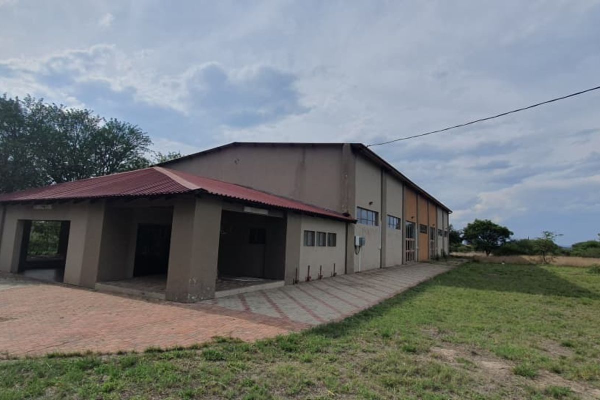 Rear view of a large industrial-style building featuring a covered patio area, corrugated metal roofing, and extensive paved surroundings on a small holding.