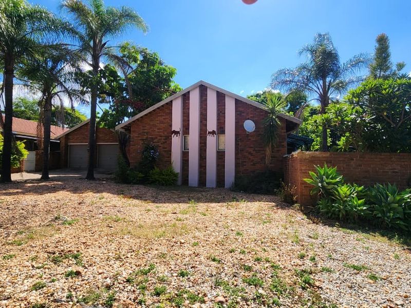Front view of a brick-faced house featuring a double garage, tiled roof, and a large unpaved driveway area surrounded by established trees and garden.