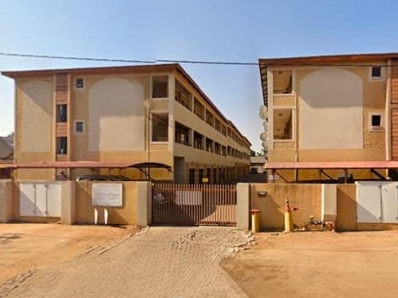 Front exterior view of a multi-story apartment complex featuring a gated entrance, paved driveway, and covered carport parking facilities.
