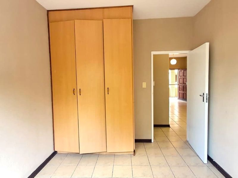 A clean, empty bedroom featuring neutral walls, tiled flooring, and a functional built-in wooden wardrobe, with a doorway leading to the adjacent living area.