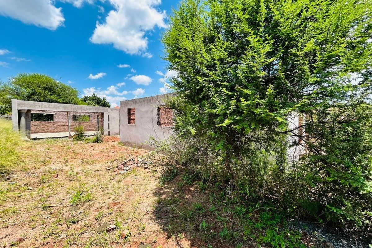 A view of an unfinished residential structure on a vacant plot, featuring exposed masonry walls and concrete framing amidst overgrown vegetation.
