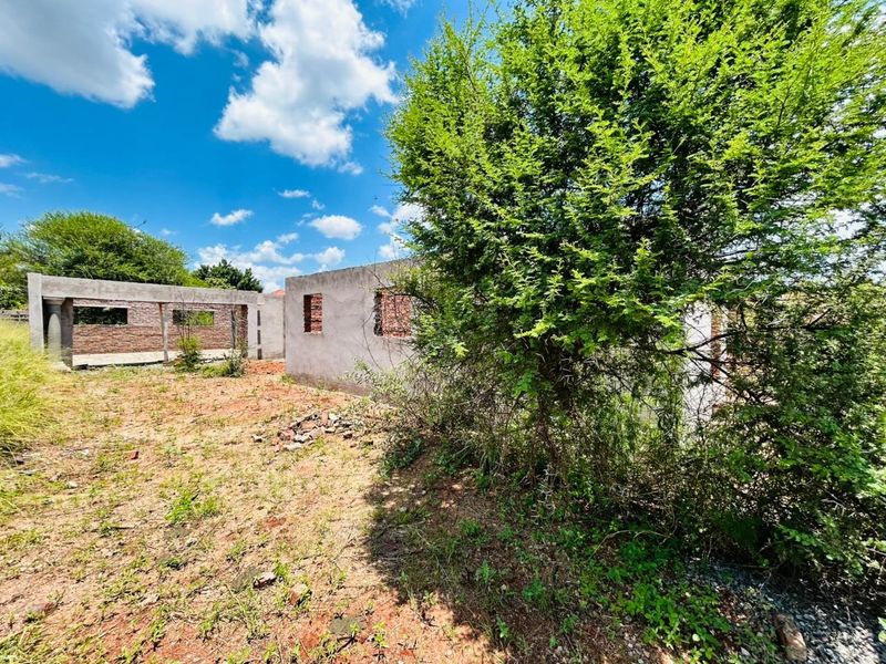 A view of an unfinished residential structure on a vacant plot, featuring exposed masonry walls and concrete framing amidst overgrown vegetation.
