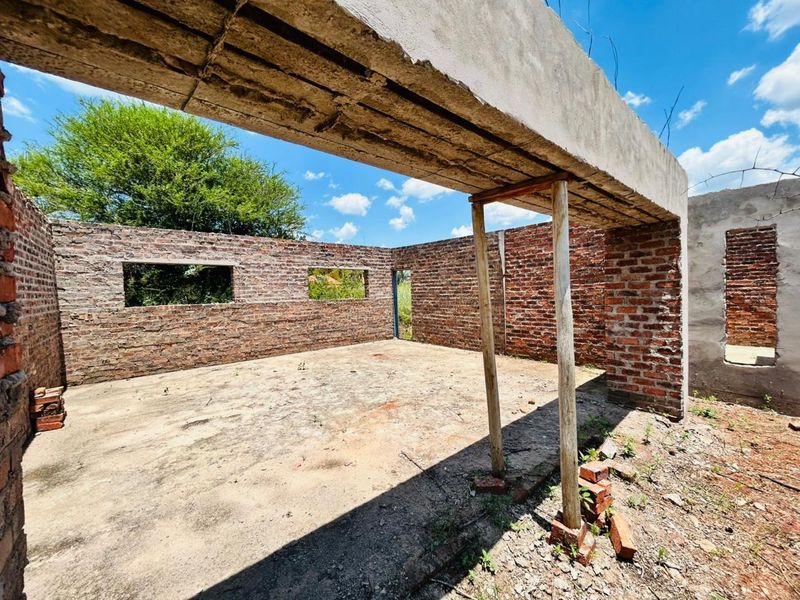 An unfinished residential structure featuring exposed brick walls and a concrete lintel, currently in a shell state on a vacant residential plot.