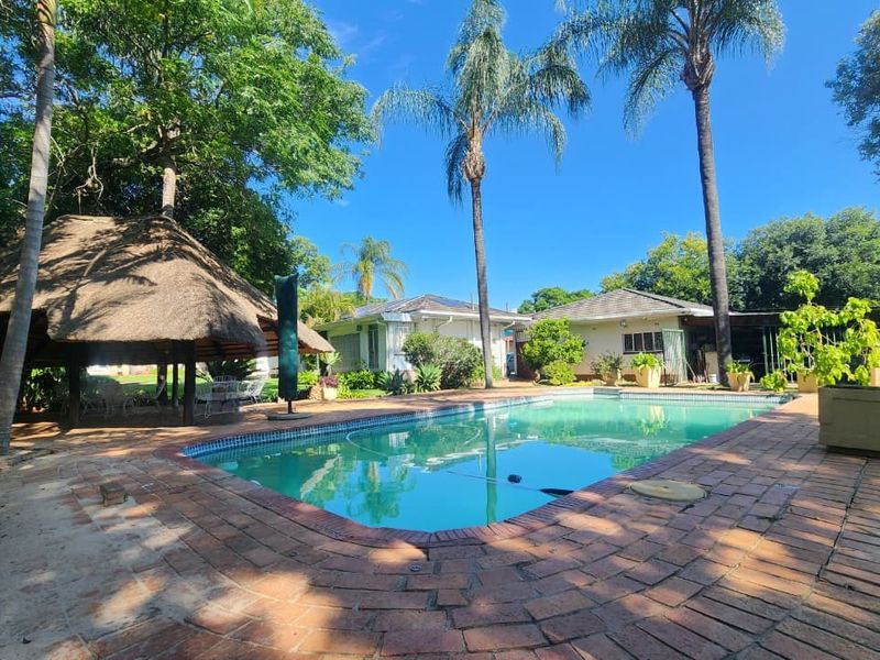 Outdoor pool area featuring a swimming pool, brick paved surround, and a traditional thatched lapa set within a landscaped garden.