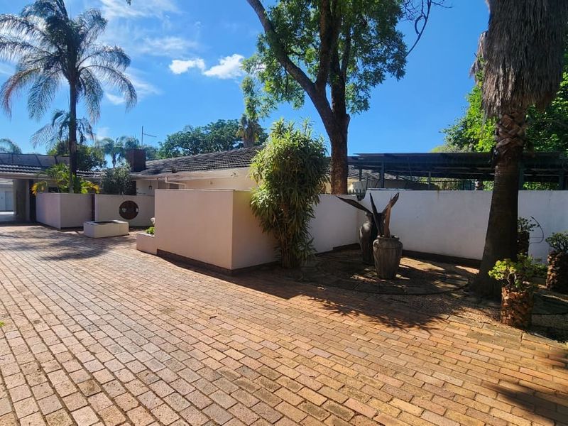 A spacious front exterior featuring a brick-paved driveway, boundary walls, and established trees leading to a covered carport area.