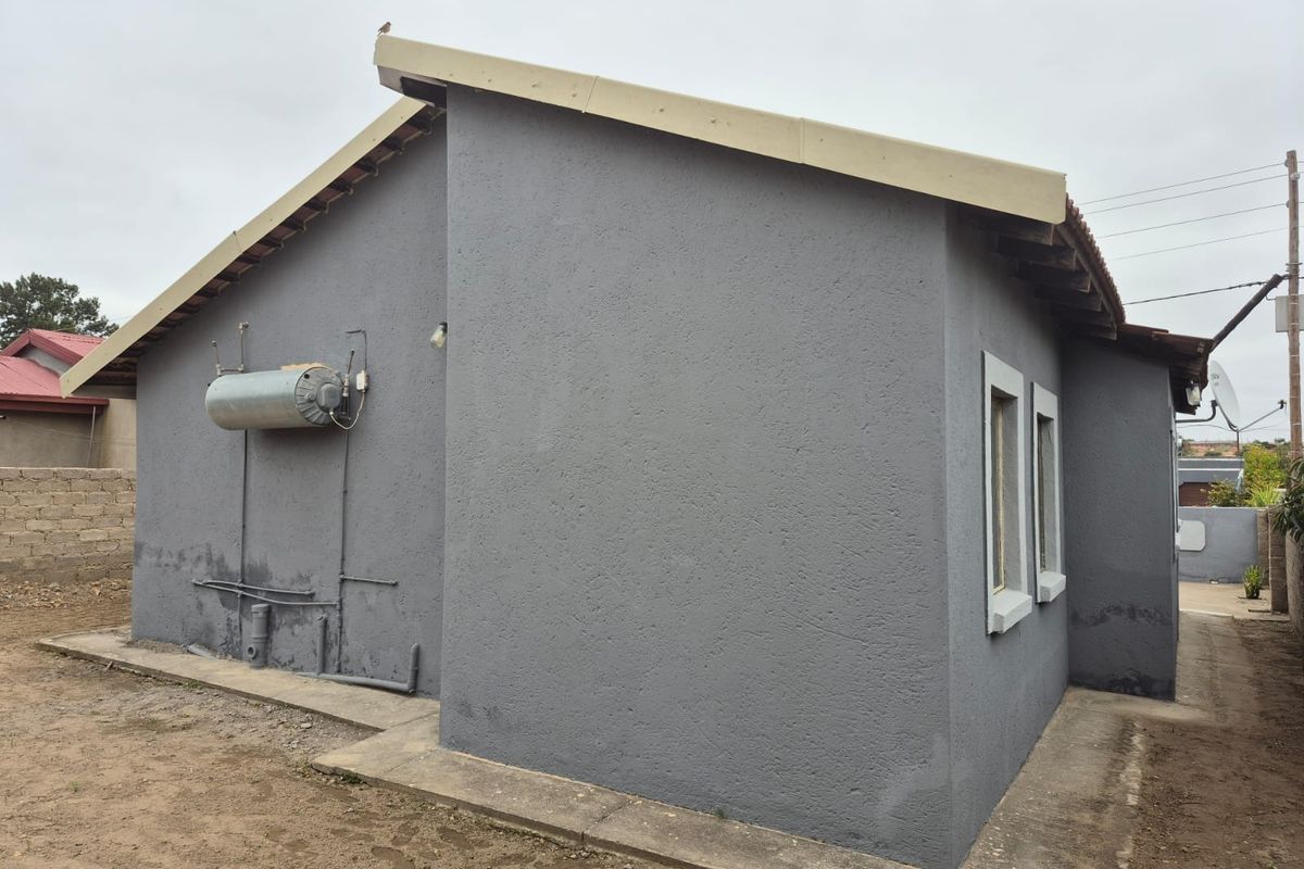 Rear exterior view of the property featuring a plastered facade, external geyser, and concrete apron with visible signs of damp on the lower wall.