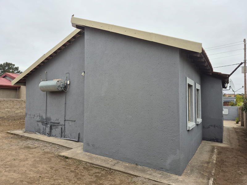 Rear exterior view of the property featuring a plastered facade, external geyser, and concrete apron with visible signs of damp on the lower wall.