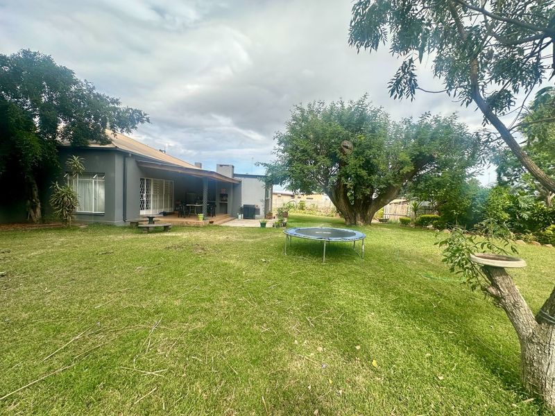 A spacious rear garden featuring a large lawn, mature trees, and a covered patio area with a built-in braai.