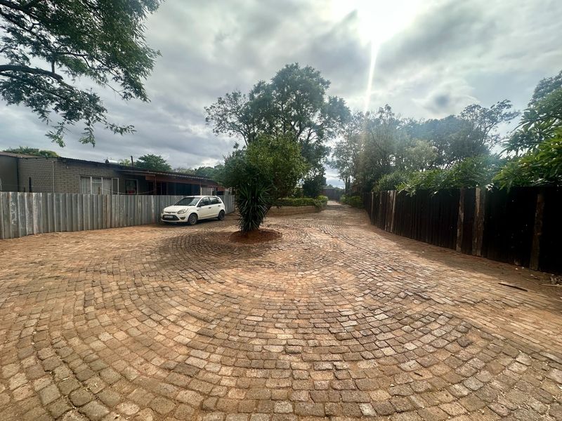 A wide driveway featuring circular patterned cobblestone paving, enclosed by a mix of corrugated metal and timber fencing with established trees.