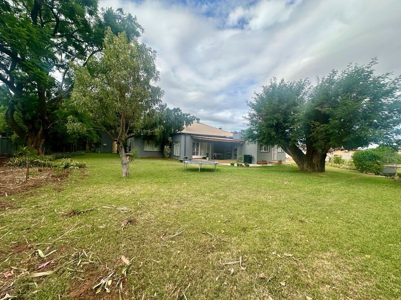 A spacious rear garden featuring a large lawn, mature trees, and a covered patio area attached to the main house structure.
