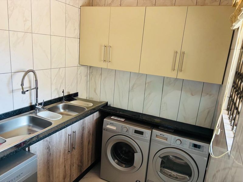A functional laundry room featuring granite countertops, double stainless steel sinks, modern cabinetry, and marble-look wall tiling.