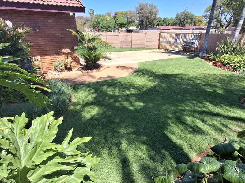 A well-maintained garden area featuring a lush lawn, established landscaping, paved driveway access, and secure perimeter walling.