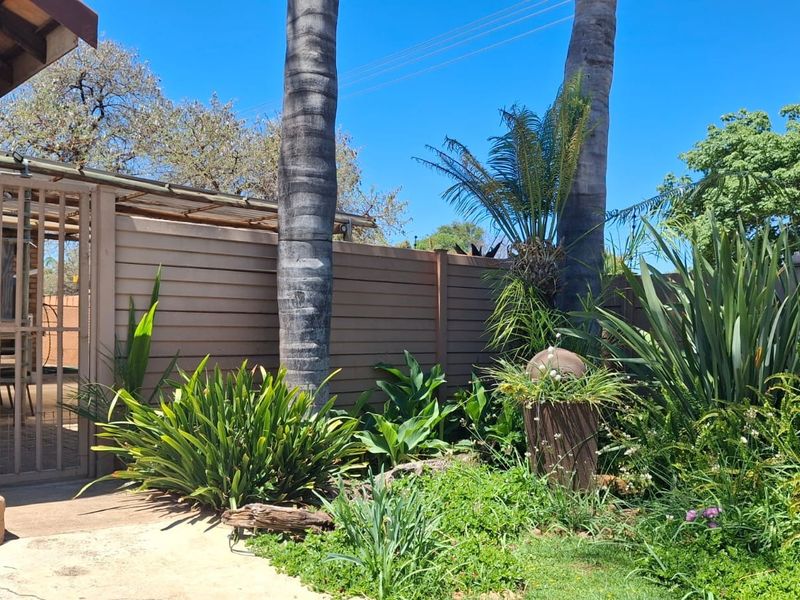 A well-established garden area featuring mature trees, lush greenery, a brick exterior wall, and a secure metal gate leading to a paved patio space.