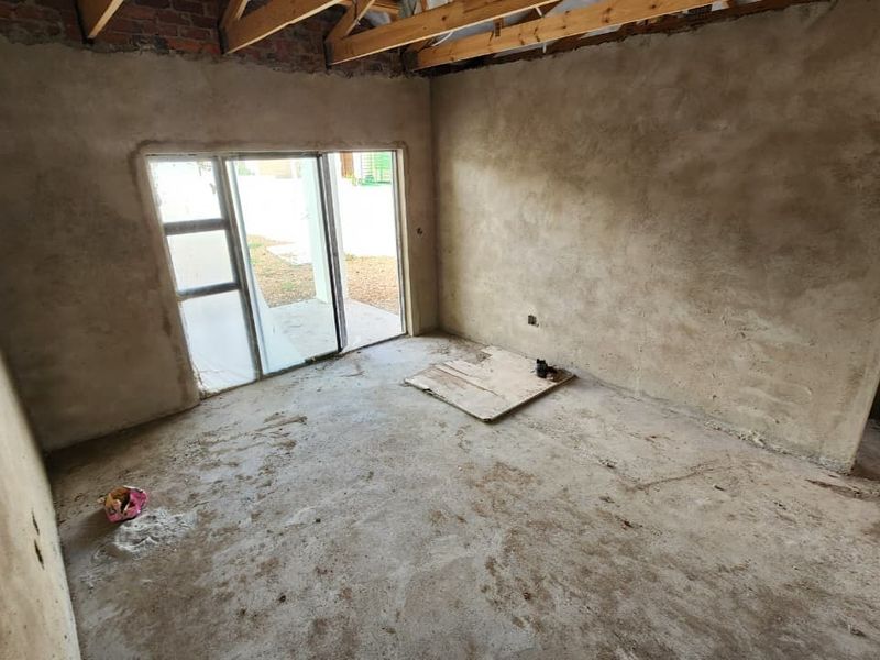 An unfinished living room space featuring exposed timber roof trusses, plastered walls, cement screed flooring, and a sliding glass door leading to the exterior.