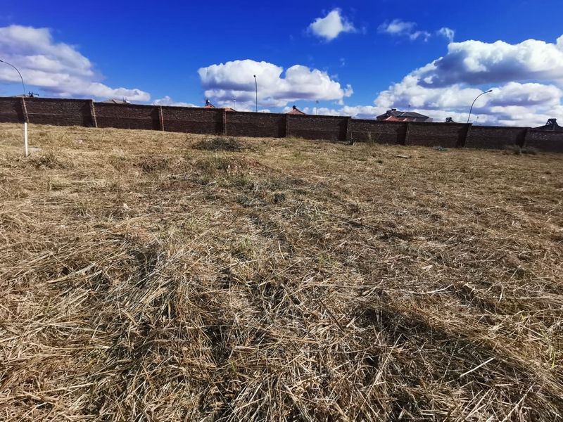 A vacant residential plot featuring dry grass and a perimeter brick boundary wall.