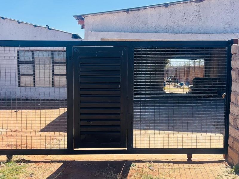 Front view of the property featuring a black metal security gate with integrated pedestrian access, flanked by brick pillars and wire mesh fencing.