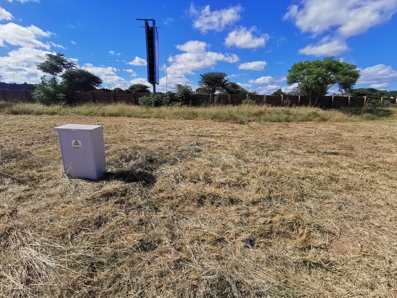 A vacant residential plot featuring dry grass, a perimeter brick wall, and an on-site electrical distribution box.