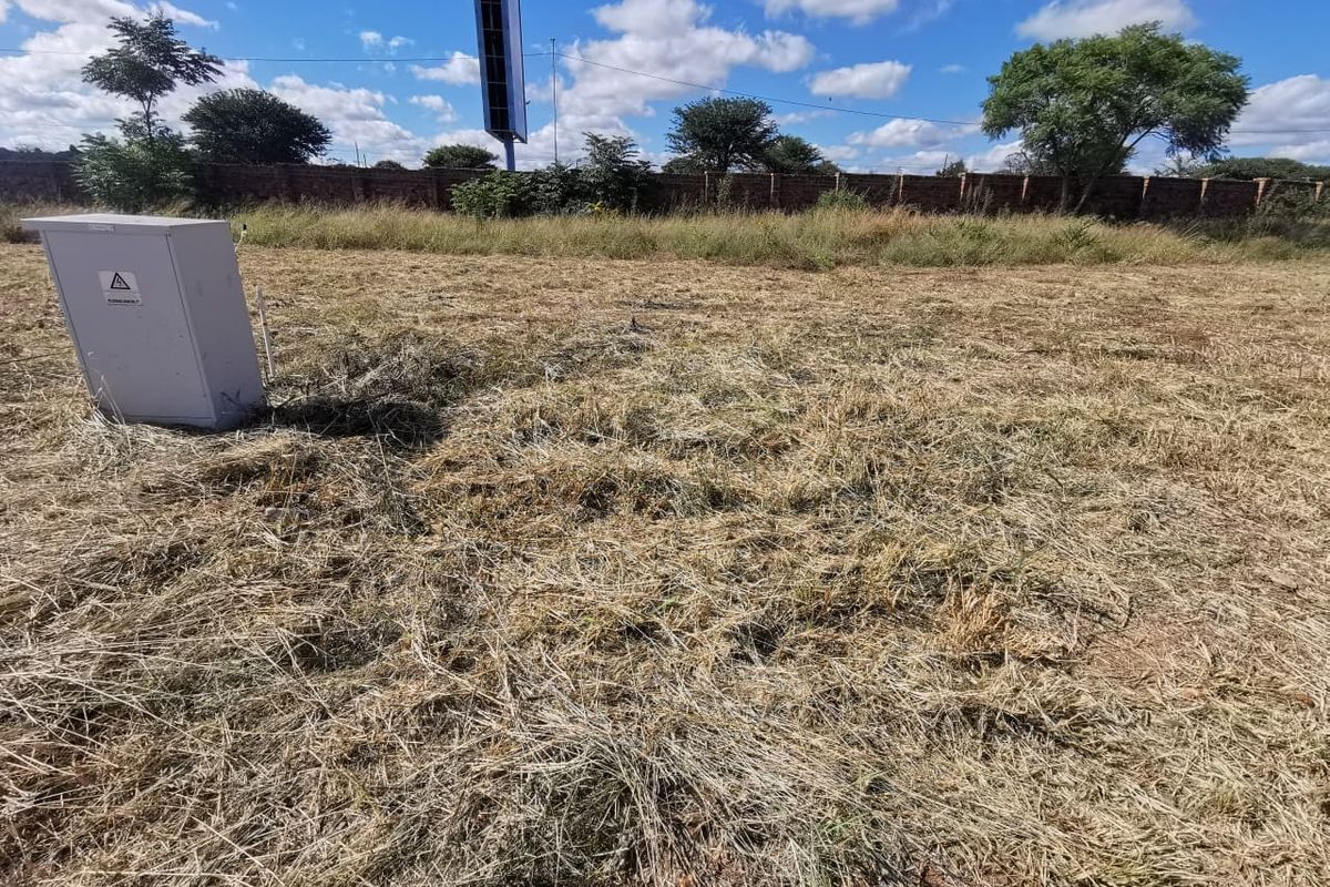 A vacant residential plot featuring dry grass, a perimeter brick wall, and an on-site electrical distribution box.