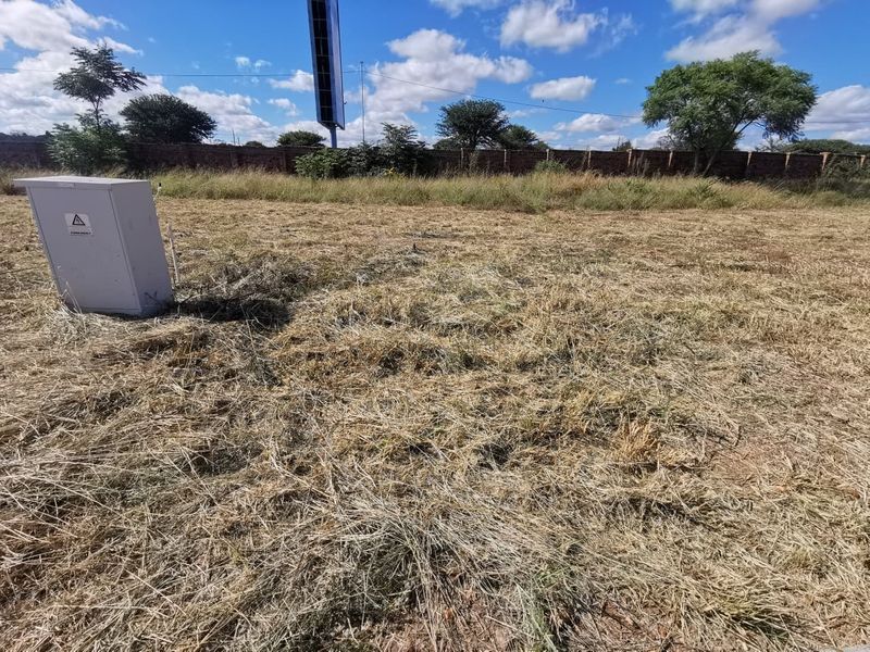A vacant residential plot featuring dry grass, a perimeter brick wall, and an on-site electrical distribution box.