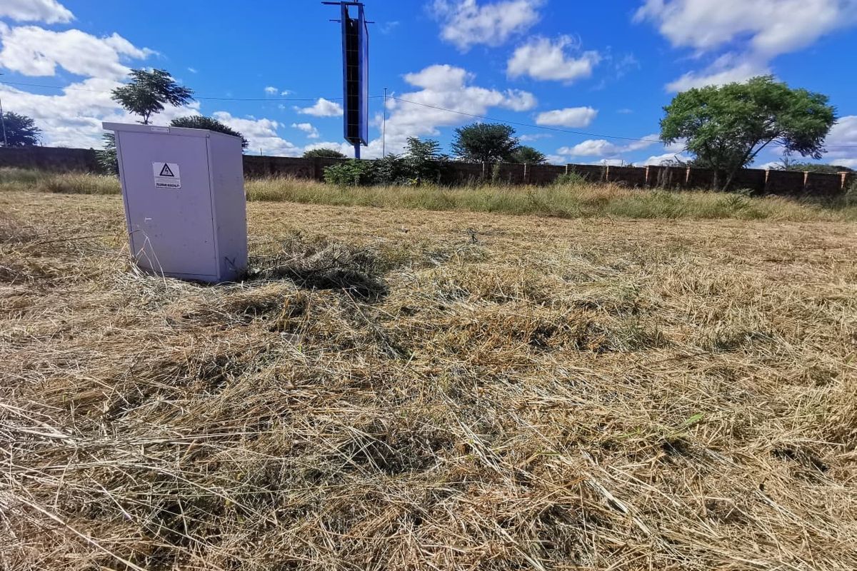 A vacant residential plot featuring dry grass, a perimeter wall, and an electrical distribution box on site.