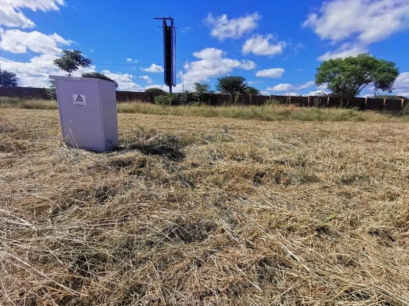 A vacant residential plot featuring dry grass, a perimeter wall, and an electrical distribution box on site.