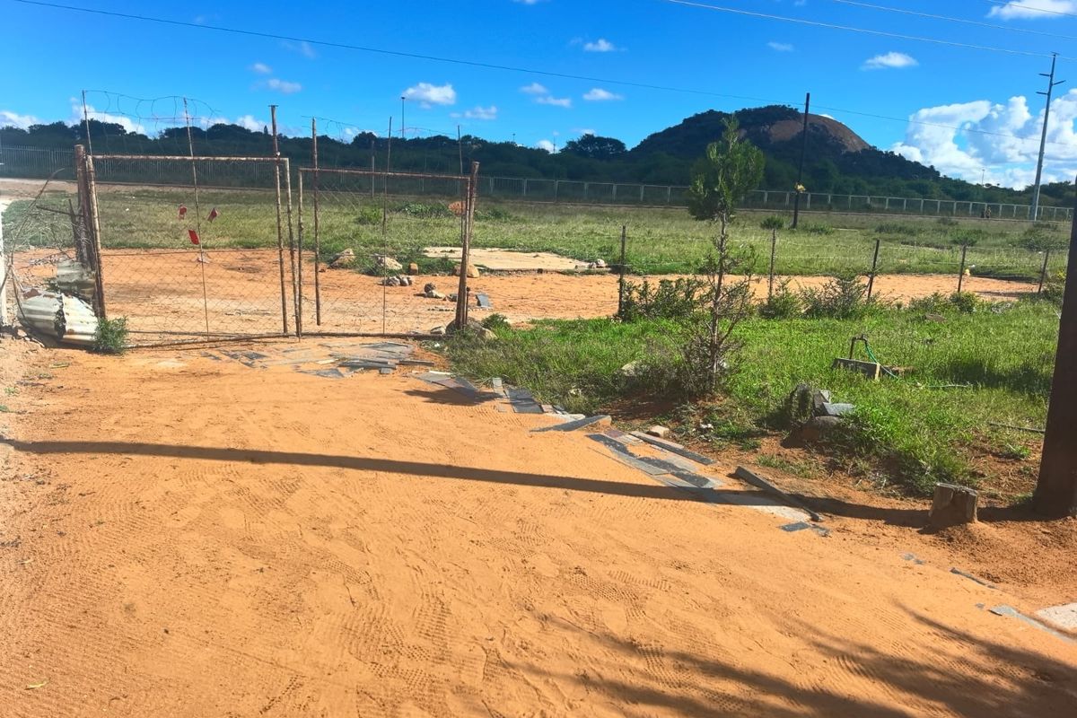 Vacant residential land featuring a metal gate, wire fencing, and an unpaved driveway with mountain views in the background.