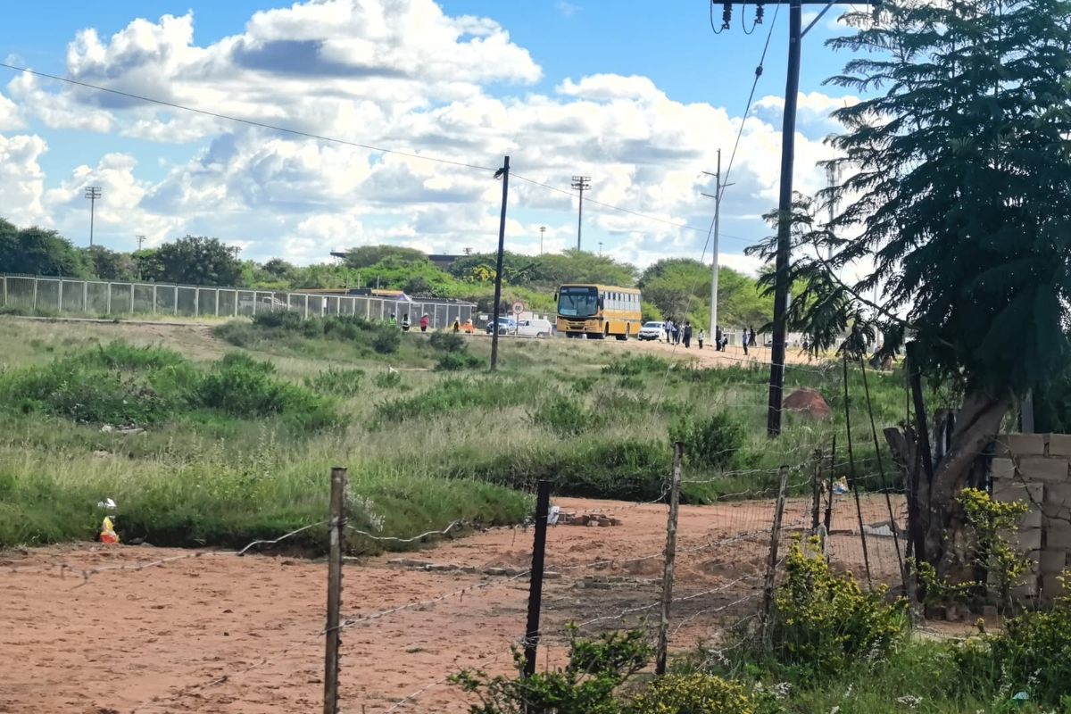 A view of a vacant residential plot featuring a barbed wire fence and utility infrastructure, with a road and bus visible in the background.