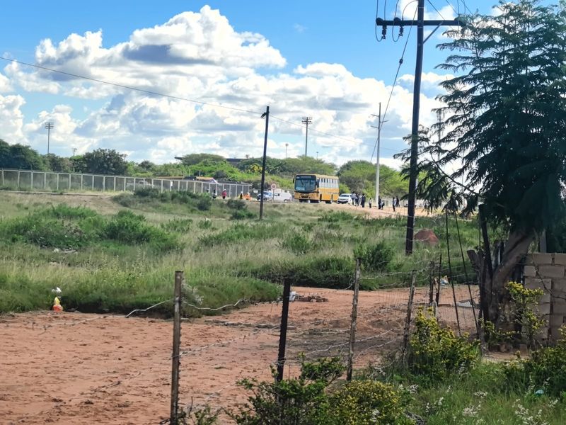 A view of a vacant residential plot featuring a barbed wire fence and utility infrastructure, with a road and bus visible in the background.