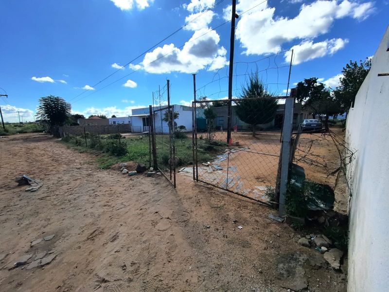 Front view of a residential plot featuring a wire mesh fence, metal gate, and unpaved access area with visible signs of wear.