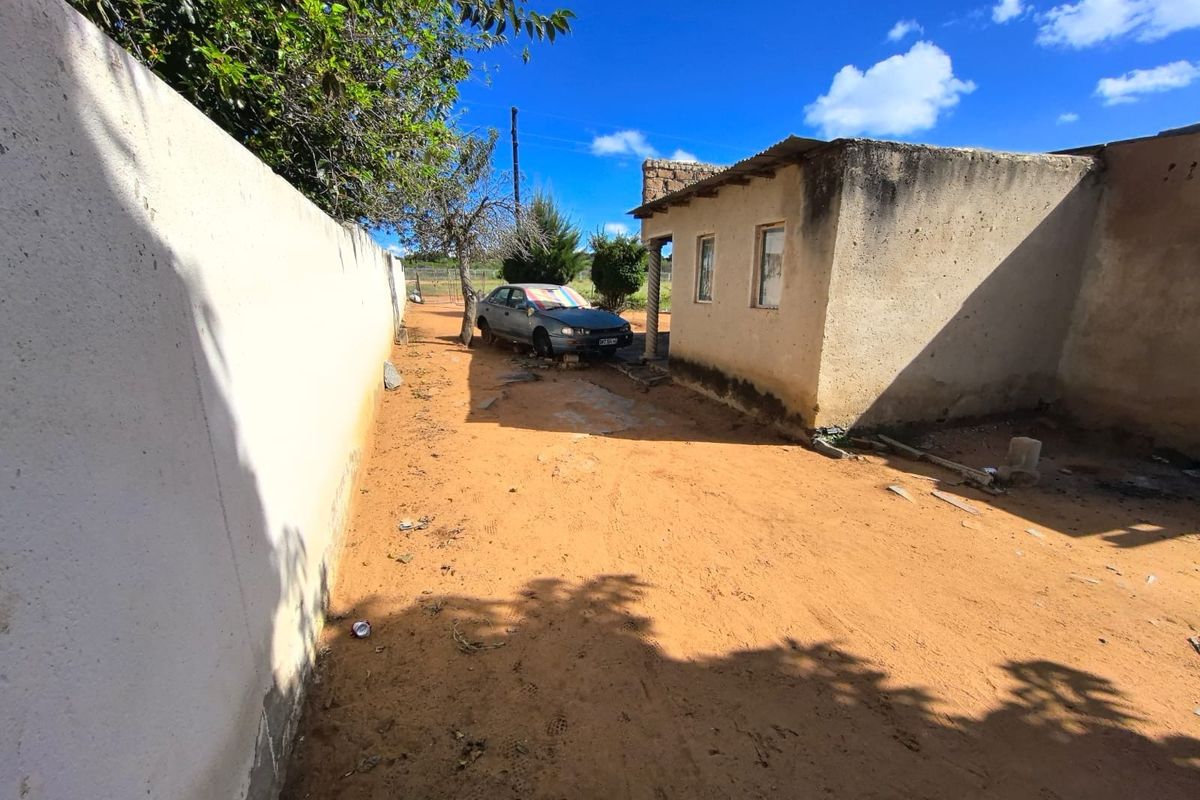 Rear exterior view of the property showing a basic structure with a corrugated metal roof and a surrounding boundary wall on an unpaved lot.
