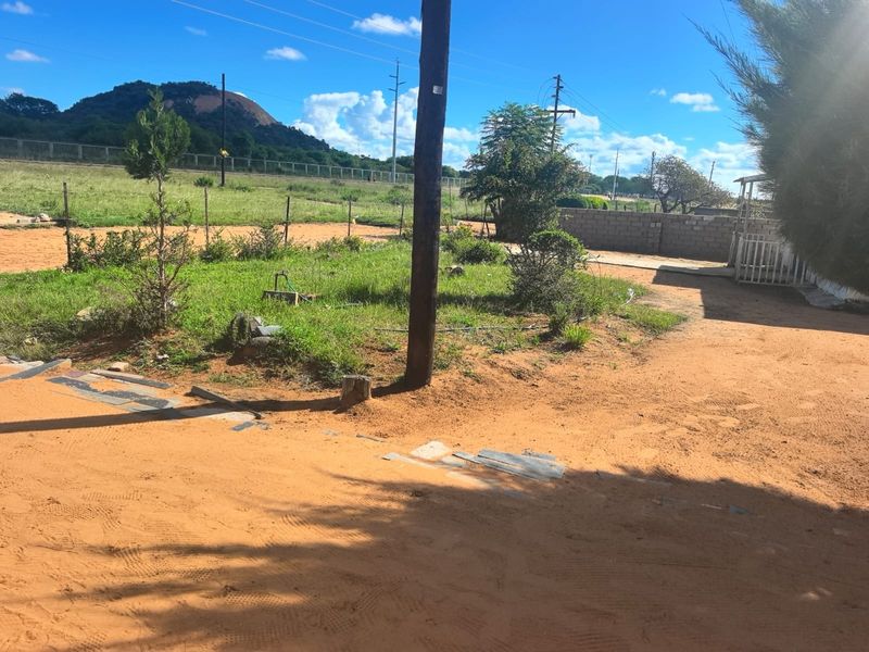 A view of a residential plot featuring a grassy area, sandy ground, and boundary fencing with a distant mountain backdrop.