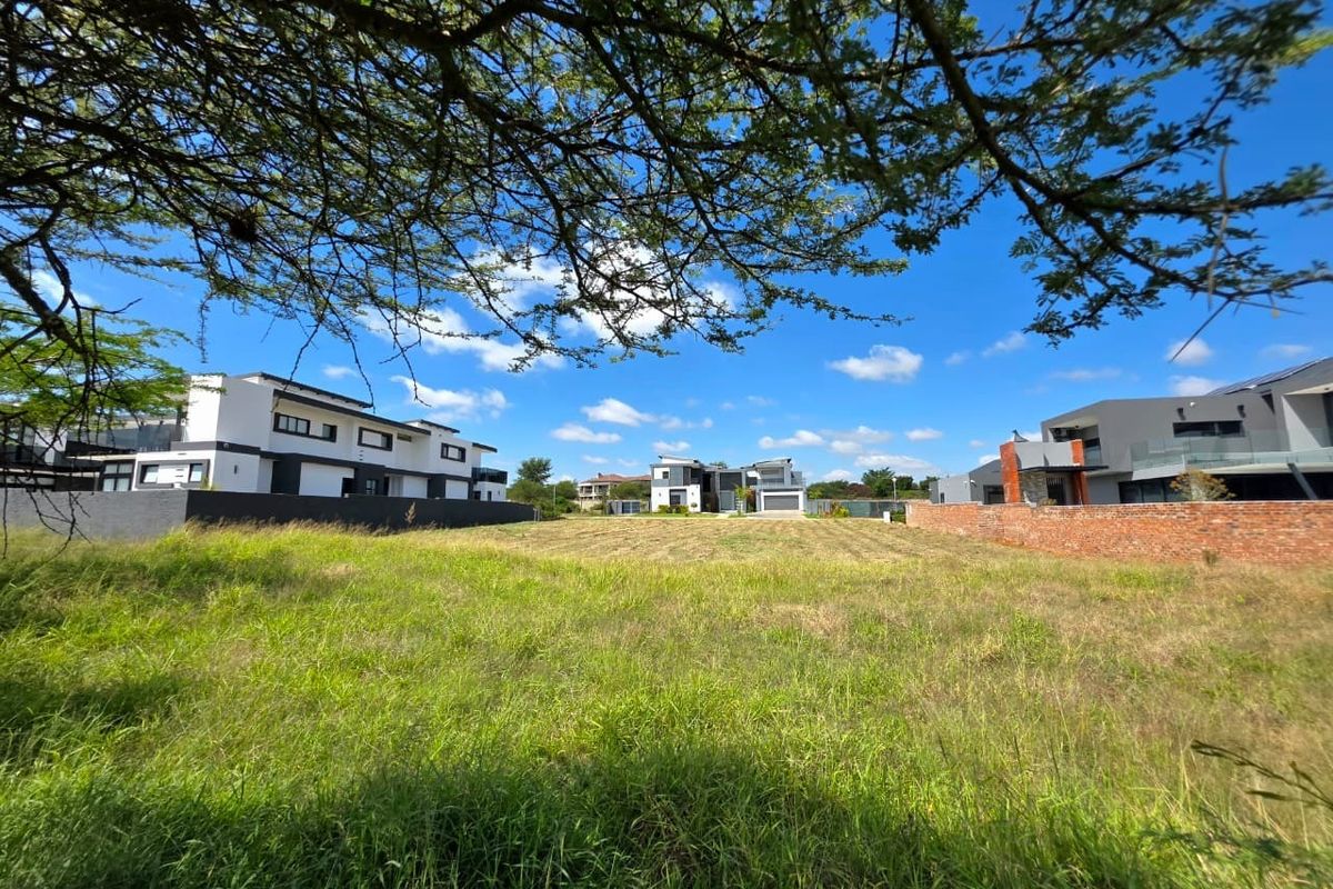 A vacant residential plot of land featuring grassed terrain, enclosed by a brick boundary wall, situated within a developed residential estate.