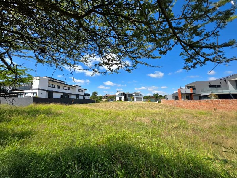 A vacant residential plot of land featuring grassed terrain, enclosed by a brick boundary wall, situated within a developed residential estate.