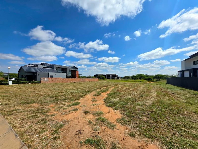 A vacant residential plot featuring a brick boundary wall, situated in a developed estate with surrounding modern homes.