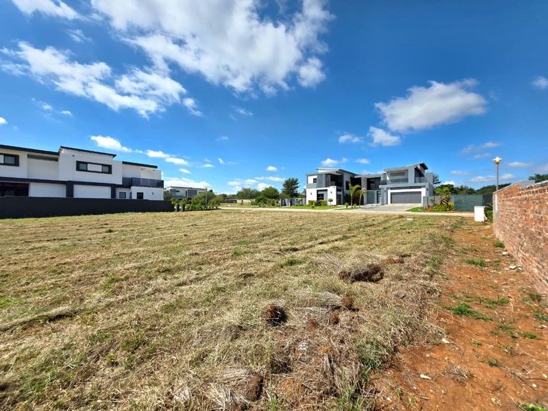 A vacant residential plot of land situated in a developed estate, featuring a brick boundary wall and clear views of neighboring modern homes.