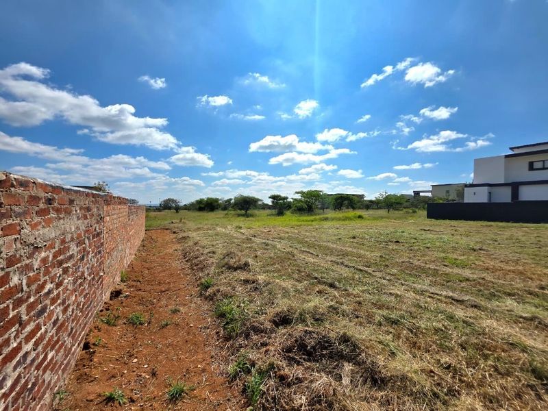 A vacant residential plot featuring a long brick boundary wall and open grassed land under a clear blue sky.