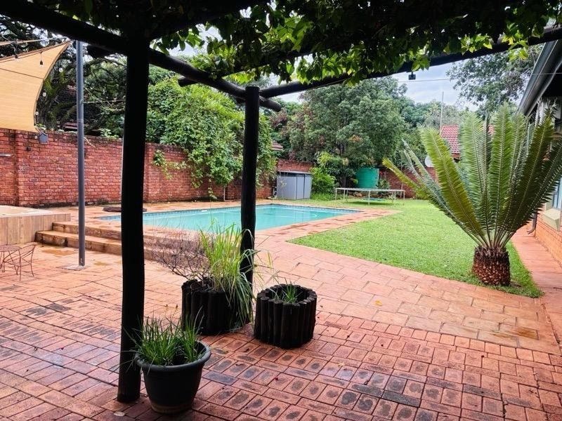 A spacious backyard featuring a swimming pool, brick-paved patio area, established lawn, and a timber pergola, enclosed by a brick boundary wall.