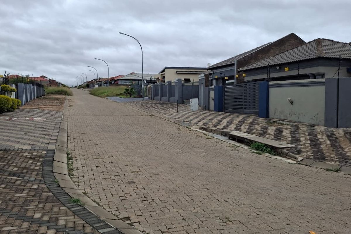 A view of a residential street featuring paved roads, sidewalks, and modern perimeter walls with electric fencing in a suburban estate.