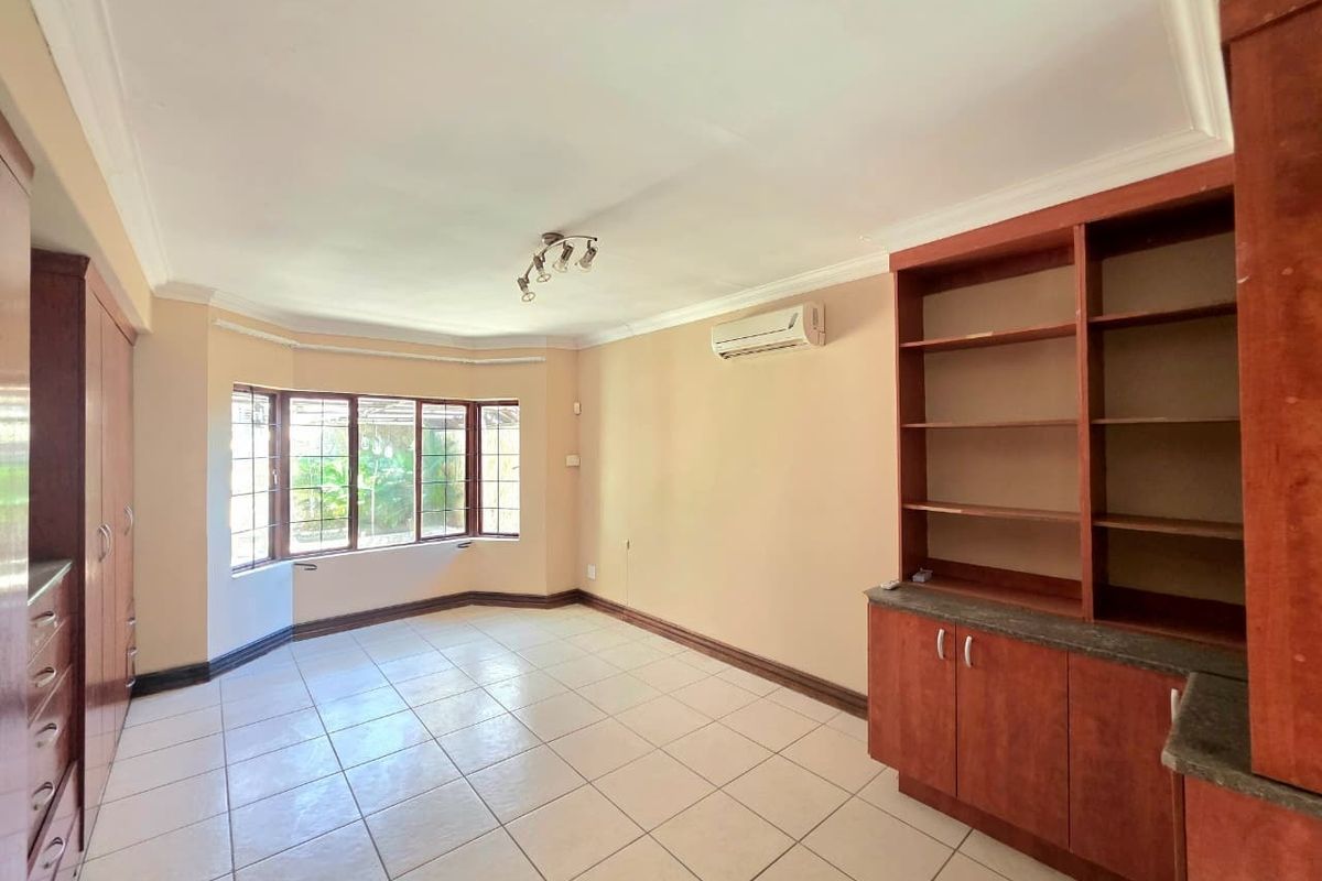 Spacious living area featuring tiled flooring, a bay window with security bars, built-in wooden cabinetry, and a wall-mounted air conditioning unit.