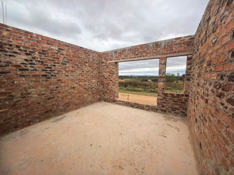 An unfinished room under construction featuring exposed brick walls and a concrete floor, offering a view of the surrounding landscape.