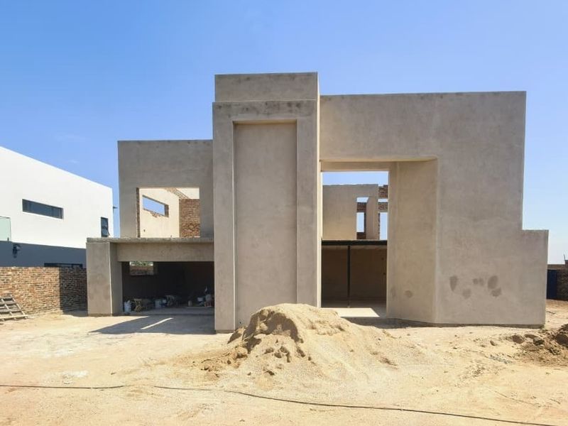 A modern architectural house under construction featuring a plastered facade, integrated garage space, and surrounding brick boundary walls on an unlandscaped plot.