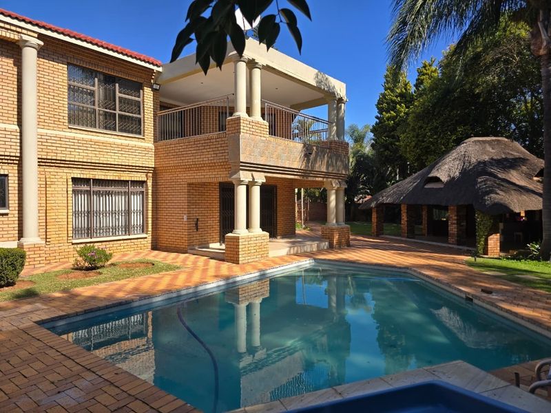 Spacious outdoor entertainment area featuring a swimming pool, paved patio, covered balcony, and a traditional thatched lapa in a landscaped garden setting.