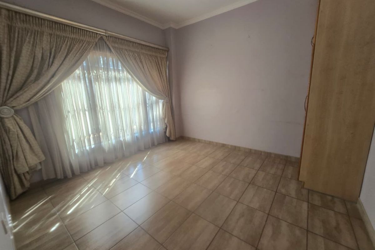 A spacious, empty bedroom featuring neutral tiled flooring, built-in wooden cabinetry, and large windows dressed with classic curtain drapes.