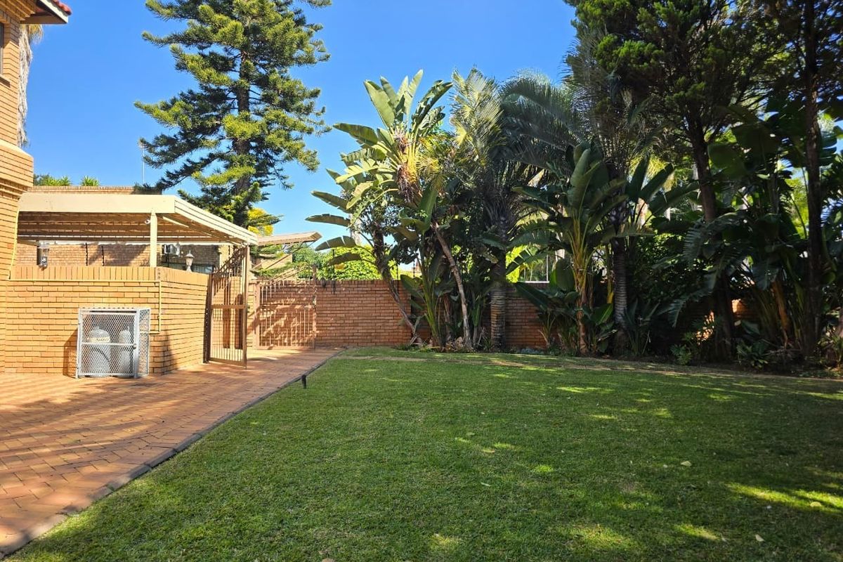 A well-maintained rear garden featuring a paved patio area, established trees, and a brick boundary wall.