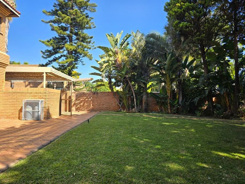 A well-maintained rear garden featuring a paved patio area, established trees, and a brick boundary wall.