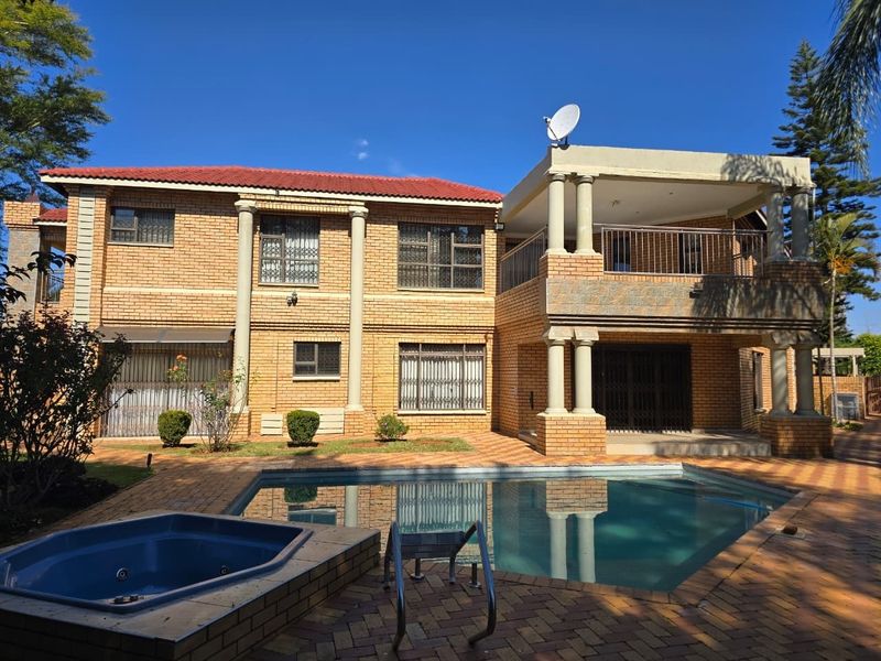 Rear view of a double-story brick house featuring a swimming pool, built-in jacuzzi, paved patio area, and an upper-level balcony with columns.
