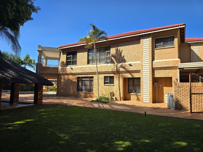 Rear view of a double-story face brick house featuring a paved patio area, a thatched lapa, and a well-maintained garden.