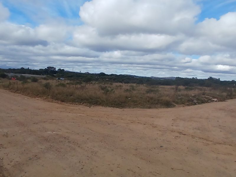 A view of a vacant residential plot situated at a dirt road intersection, featuring natural vegetation and perimeter fencing.