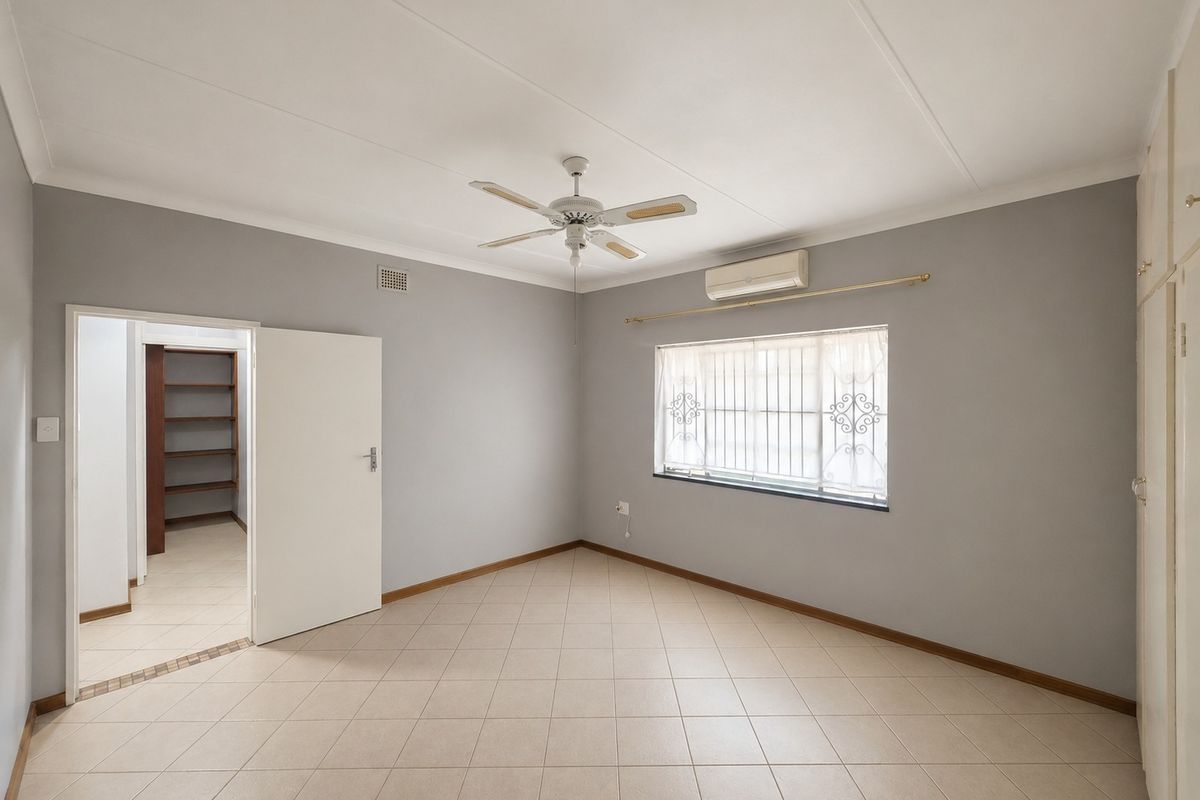A spacious, empty bedroom featuring neutral grey walls, tiled flooring, a ceiling fan, air conditioning unit, and built-in storage cupboards.