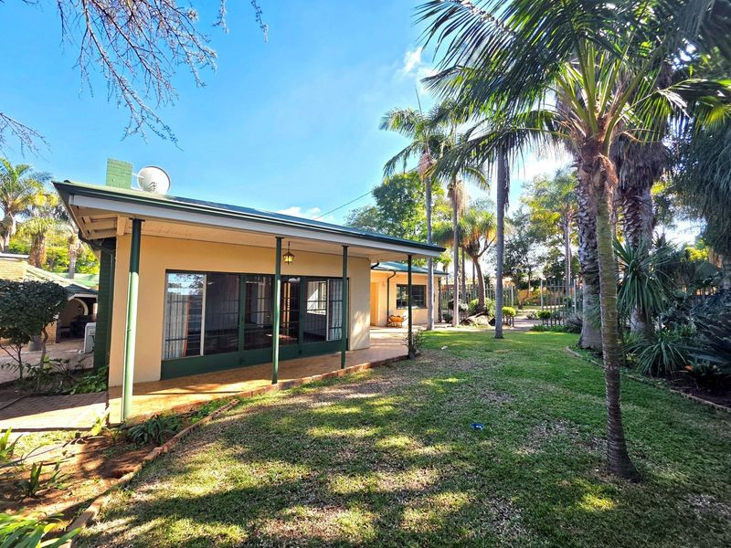 Rear view of the house featuring a covered patio with brick paving, sliding glass doors, and a well-maintained garden with mature palm trees.
